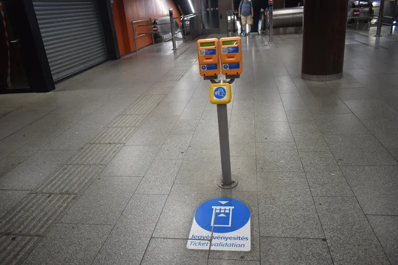 Corvin-negyed駅の刻印機。改札機は無く、紙の券はオレンジの機械の差込口へ入れる。下の黄色い面はスマホをかざすNFC面で、床の青い丸「Jegyérvényesítés／Ticket validation」が刻印の位置を示す